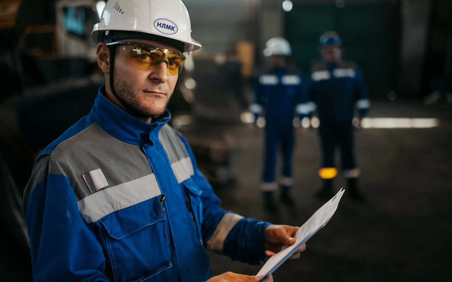 Steel-factory professional reviewing fabrication documents while production staff work in the background
